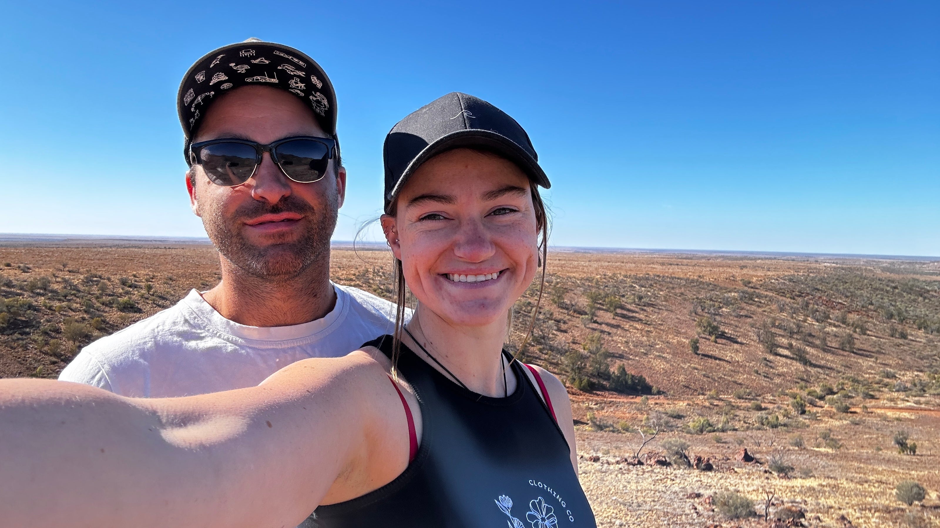 a young aussie couple wearing caps and shirts by aus wild clothing co taking a selfie with expansive backdrop of the flinders ranges in South Australia